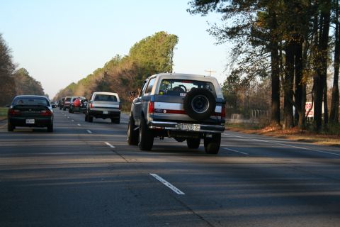 1982 Ford Bronco - A lifted, two-tone blue and white Ford Bronco with a spare tire mounted on the rear is driving down a highway. Several other vehicles are v…