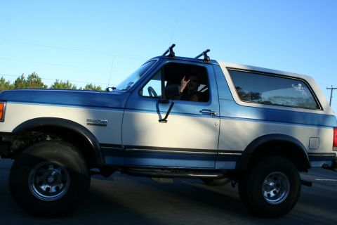 1982 Ford Bronco - A two-tone blue and white Ford Bronco is driven on a road under a clear sky.