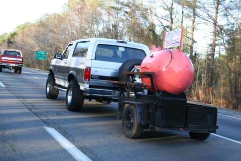 1982 Ford Bronco - A silver 1982 Ford Bronco tows a large pink smoker trailer down the highway, with another truck visible in the background.