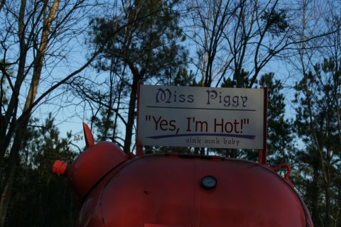 1982 Ford Bronco - A large, red, pig-shaped smoker is displayed outdoors under a sign that reads 'Miss Piggy "Yes, I'm Hot!"'.