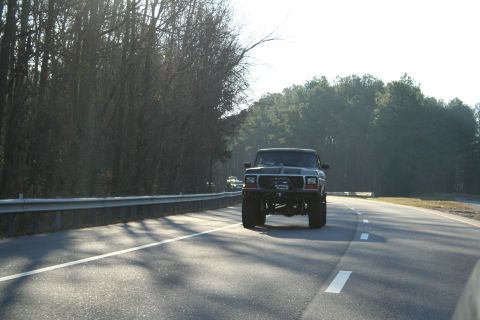 1982 Ford Bronco - A lifted black Ford Bronco is seen driving on a highway with trees in the background.