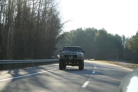 1982 Ford Bronco - A lifted black truck drives down a highway, its front grille and headlights clearly visible.