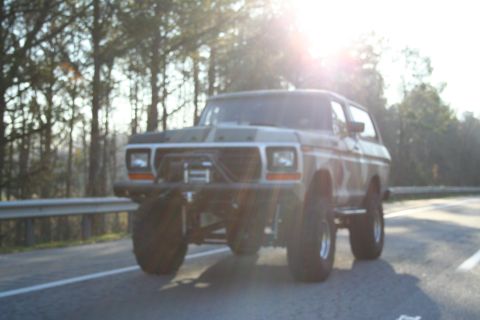 1982 Ford Bronco - This modified Ford Bronco is driving down a road with trees in the background and the sun shining.