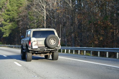 1982 Ford Bronco - Lifted 1980s Ford Bronco with spare tire on the rear is seen driving on a highway.