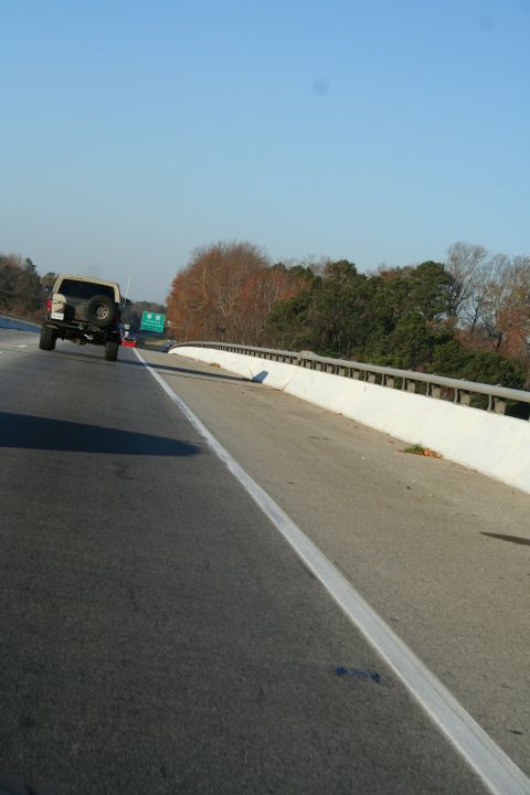1982 Ford Bronco - A tan SUV with a spare tire mounted on the rear door travels down a highway under a clear blue sky.