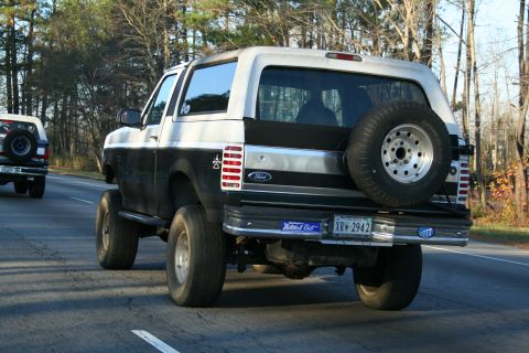1982 Ford Bronco - A black and silver Ford Bronco is seen from the rear, driving on a road with another vehicle ahead.