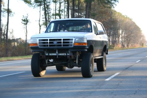 1982 Ford Bronco - This lifted two-tone SUV, likely a 1992 Ford Bronco, is captured from the front while driving on a highway.