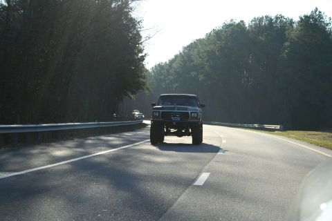 1982 Ford Bronco - Lifted Ford Bronco driving on a highway with trees lining the road.