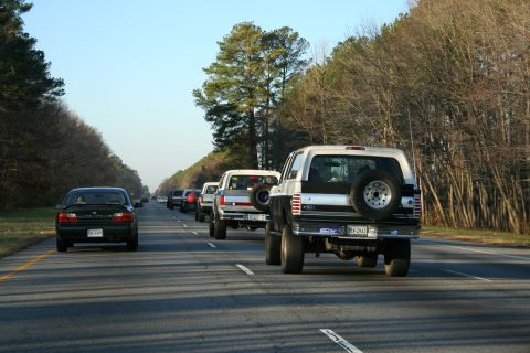 1982 Ford Bronco - A line of Ford Broncos drives down a highway lined with trees.
