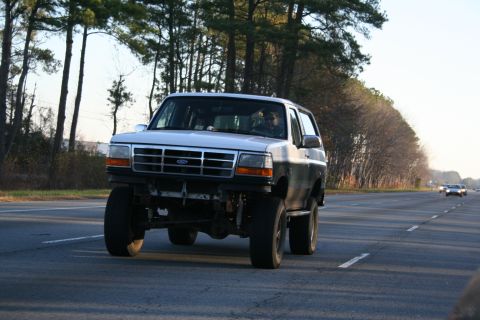 1982 Ford Bronco - This lifted Ford Bronco is driving on a highway with trees lining the road.