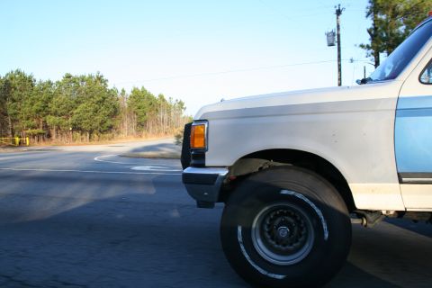 1982 Ford Bronco - Close-up view of the front corner of a pickup truck, showing its wheel and fender against a backdrop of trees.