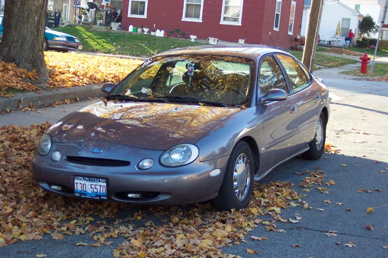 Gray Ford Taurus parked on a street covered in fallen autumn leaves. License plate reads T30 5570