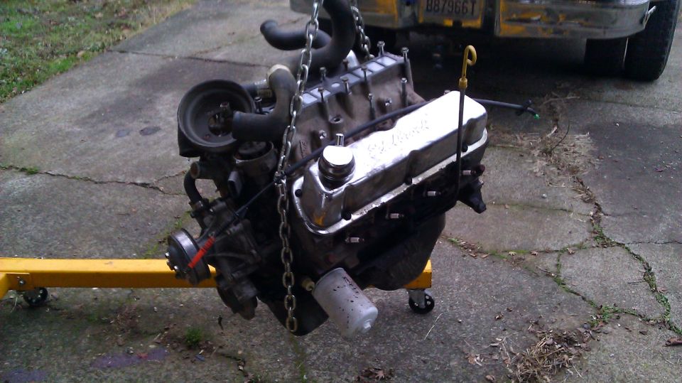V8 engine with chrome valve covers hanging from a yellow engine hoist on a concrete driveway