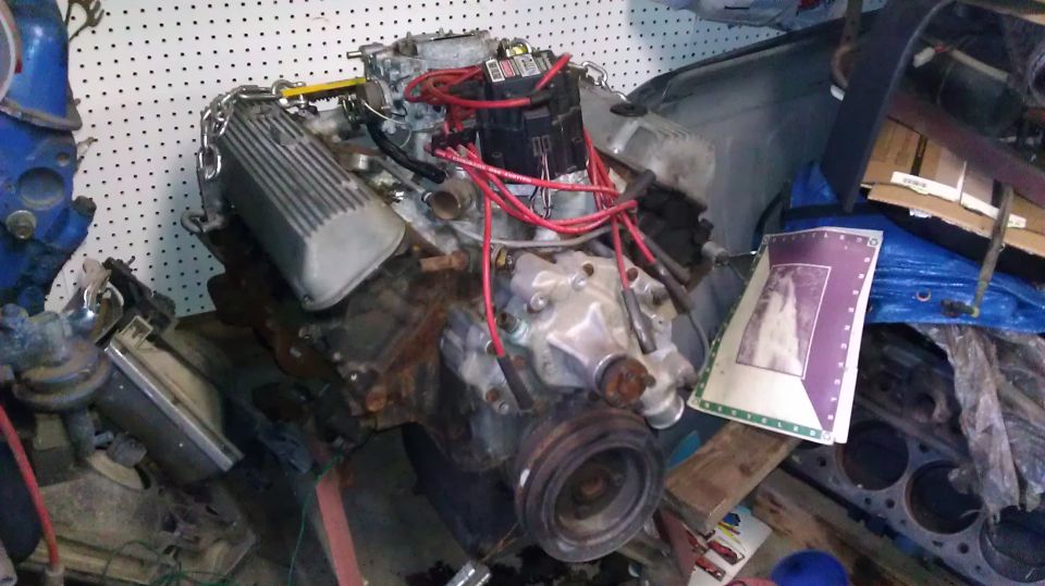 V8 engine with red spark plug wires and silver valve covers, sitting in a cluttered garage