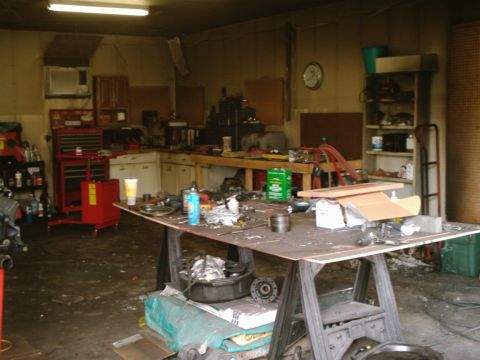 1989 Ford Bronco - A cluttered garage scene with various tools and automotive parts scattered across workbenches and the floor.