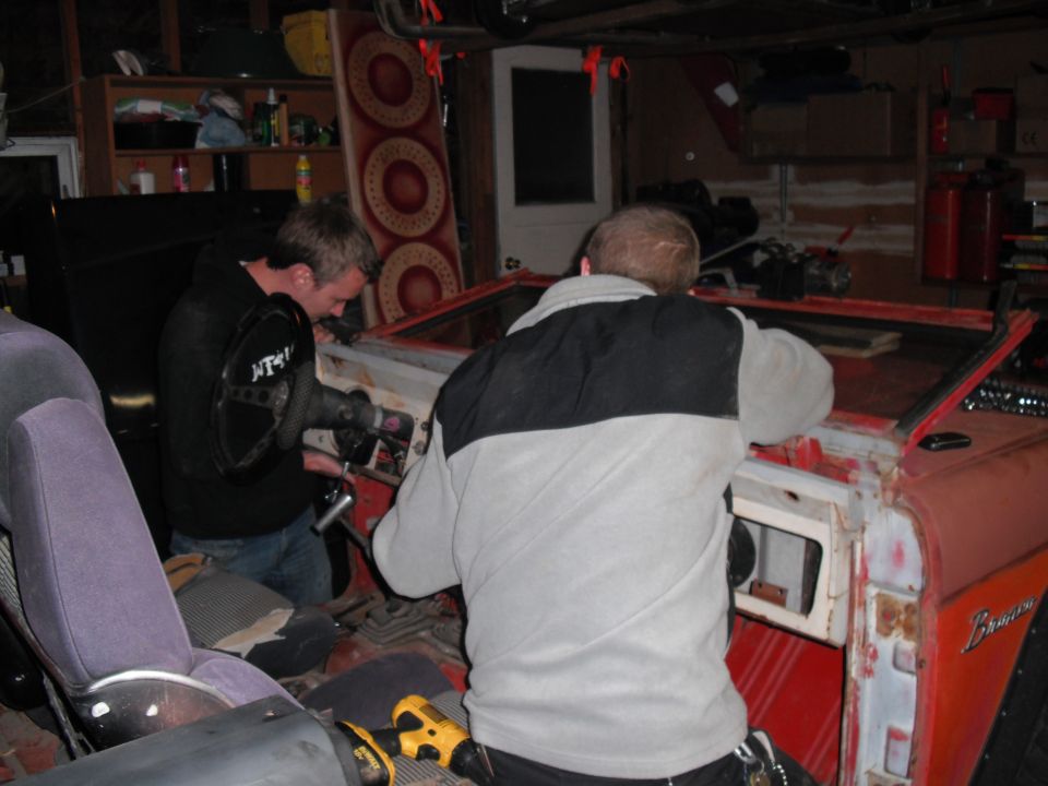 Two men working on the interior of a red 1968 Ford Bronco, one using a tool near the steering column
