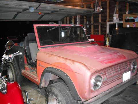 1968 Ford Bronco - A red 1968 Ford Bronco sits in a garage, showing its front passenger side with faded paint and black fender flares.