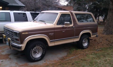 1984 Ford Bronco - This two-tone SUV is parked next to another vehicle in a driveway.
