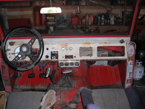 1968 Ford Bronco - Interior of a stripped early Ford Bronco looking across the white rusty dash from behind the black three-spoke steering wheel, with a bare…
