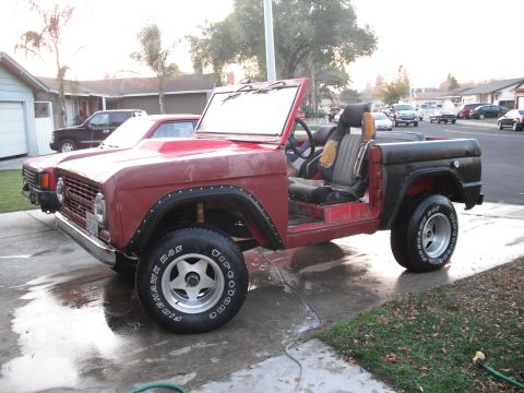 1968 Ford Bronco - Driver-side view of a red early Ford Bronco standing doorless and topless on a wet suburban driveway, hood raised, with cloth-insert bucket…