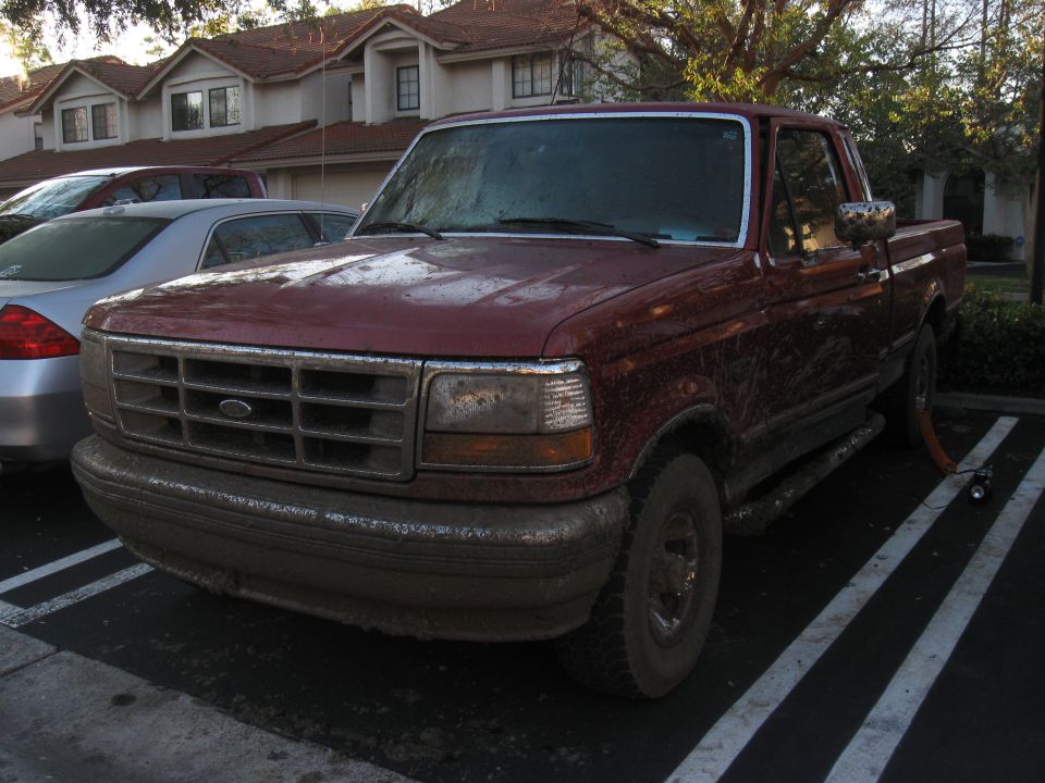 1995 Ford F-150 pickup truck, front view, covered in mud. Another car is parked to the left