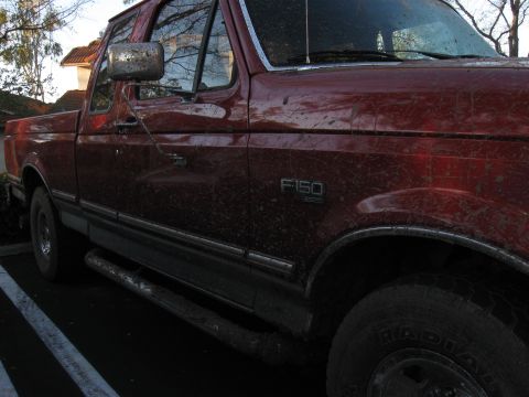 1995 Ford F-150 - A muddy red 1995 Ford F-150 pickup truck is parked, showing its side profile and off-road condition.