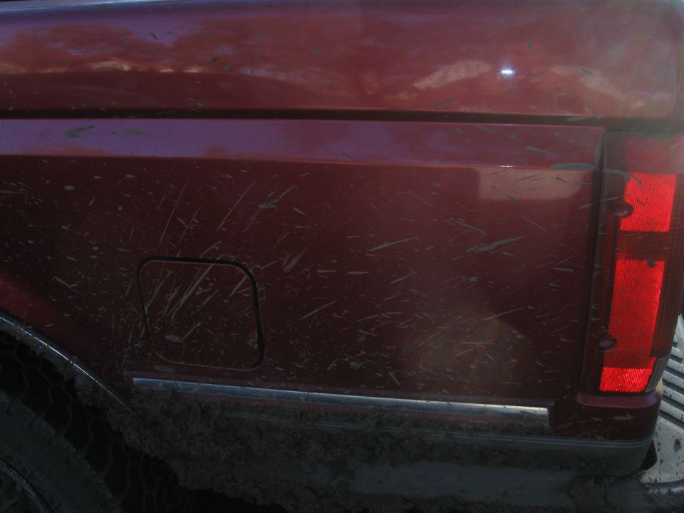 Mud-splattered red pickup truck rear side with fuel door and taillight visible. Supermotors.net logo at bottom right