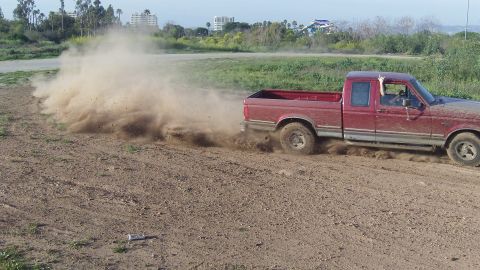 1995 Ford F-150 - A red pickup truck drifts on a dirt road, creating a large dust cloud. The vehicle's body style suggests a model year around 1992.