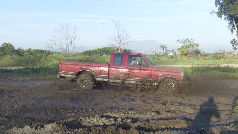 1995 Ford F-150 - A red pickup truck navigates a muddy terrain, kicking up dirt as it drives through the mire.
