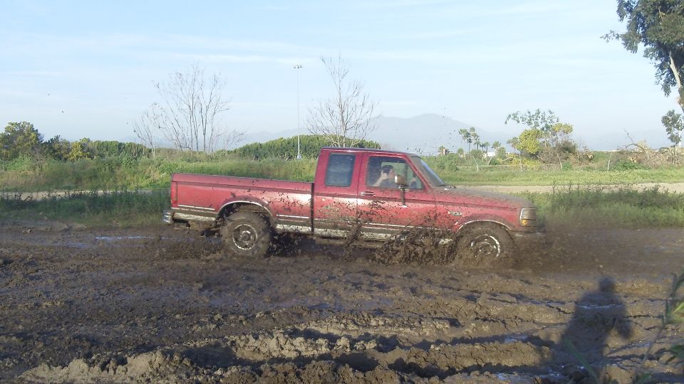 Red pickup truck with a crew cab driving through mud, splashing dirt. The truck appears to be from the early to mid-1990s