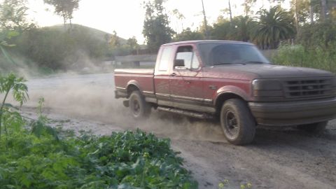 1995 Ford F-150 - A red 1995 Ford F-150 navigates a dusty dirt road, kicking up a cloud of dust as it goes.