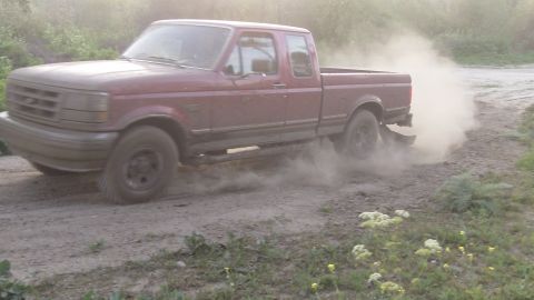 1995 Ford F-150 - A red pickup truck is driving on a dirt road, creating a cloud of dust.