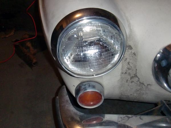Close-up of round headlight with chrome ring and amber turn signal on a dirty white vintage car front fender