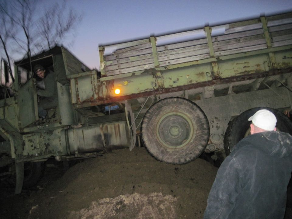 Military truck with wooden bed stuck in dirt, driver in cab, person watching