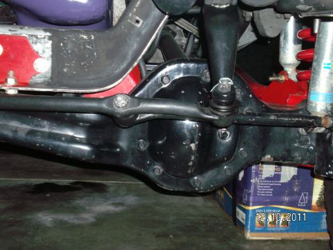 1996 Ford Bronco - Underbody view of a black solid front axle with its differential cover, showing the tie rod and drag link joints running over the housing i…
