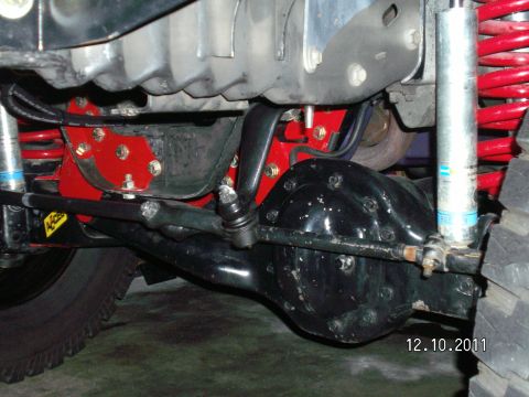 1996 Ford Bronco - Underside of a lifted 4x4 front suspension showing a black solid axle with bolted differential cover, tie rod and drag link running above t…