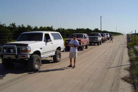 1993 Ford Bronco - Multiple Ford Broncos drive down a dusty dirt road under a clear blue sky.