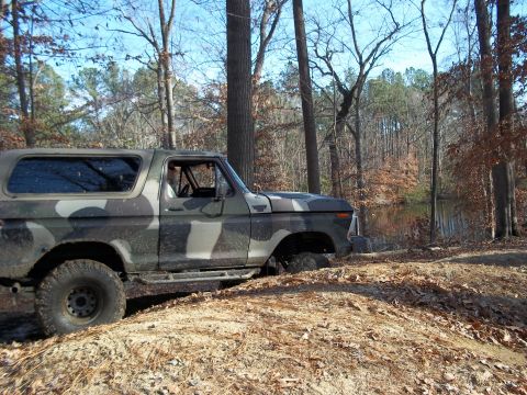 1979 Ford Bronco - A camouflaged Ford Bronco navigates rough terrain in a wooded area with a lake in the background.