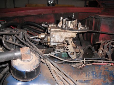 1968 Ford Bronco - Close-up of a weathered two-barrel carburetor sitting on the intake of a blue engine, air cleaner removed, with the throttle bellcrank, ret…