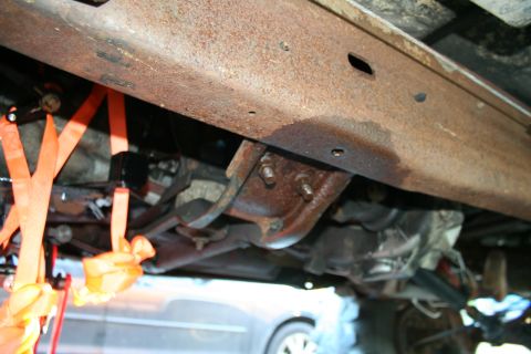 1982 Ford Bronco - undercarriage view of a rusty truck frame crossmember and leaf spring, with an orange ratchet strap and a car visible below.
