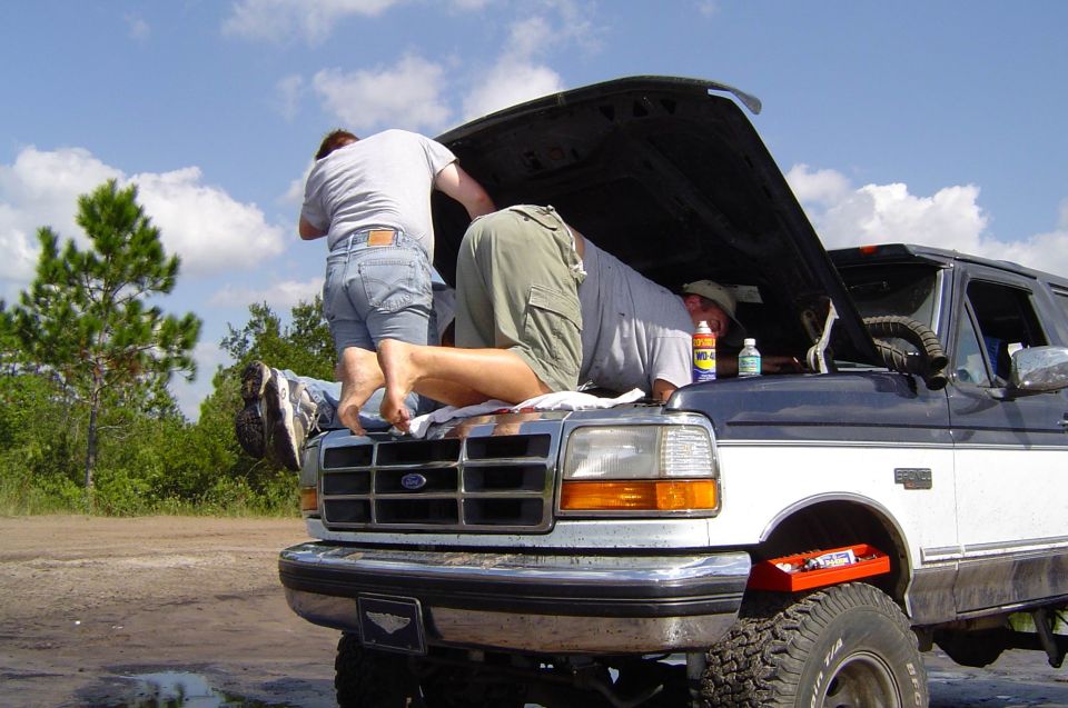 People working under the raised hood of a lifted white Ford Bronco on a sandy trail pull-off
