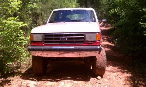 1987 Ford Bronco - Front view of a 1987 Ford Bronco driving on a dirt path through trees.