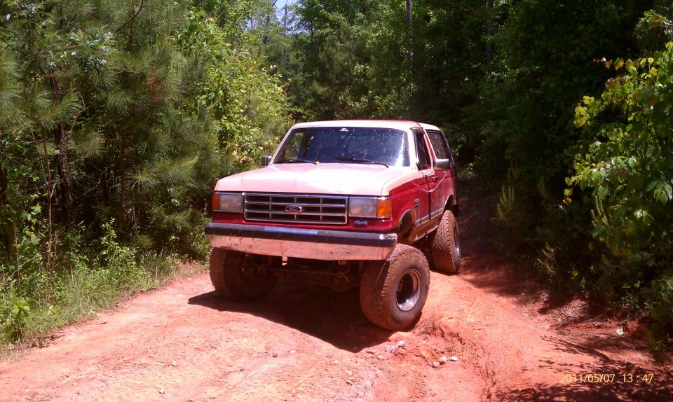 1987 Ford Bronco with large tires on a dirt trail, surrounded by trees