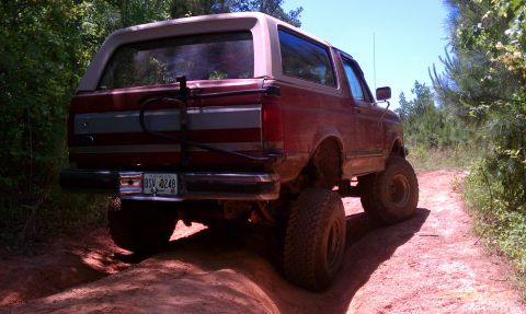 1987 Ford Bronco - A lifted red 1987 Ford Bronco navigates a dusty, wooded trail with a spare tire mounted on the back.