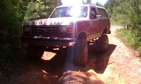 1987 Ford Bronco - A lifted 1987 Ford Bronco navigates a rough dirt path surrounded by trees.