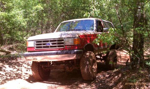 1987 Ford Bronco - A lifted 1987 Ford Bronco navigates a wooded trail with mud-splattered tires and a two-tone paint scheme.