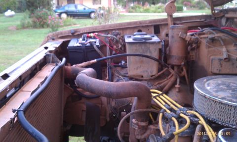 1987 Ford Bronco - The engine bay of a vehicle, with a battery and various hoses and wires visible. A car is parked in the background.