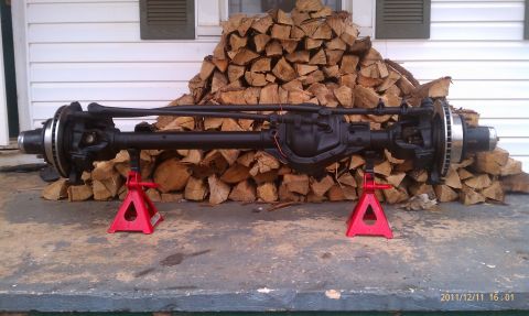 1987 Ford Bronco - Front axle assembly supported by jack stands, with a large pile of firewood in the background.