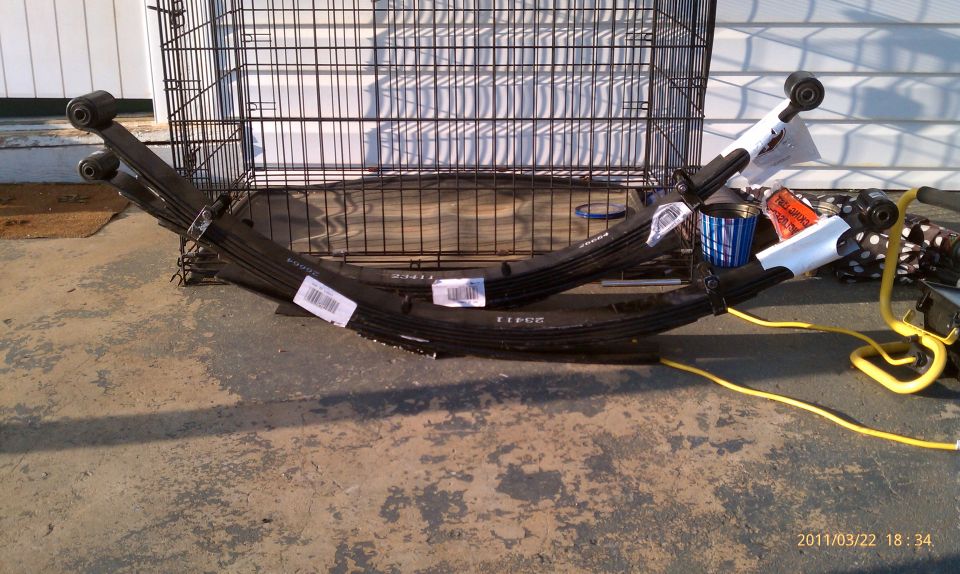 Two black multi-leaf leaf springs with barcode labels on concrete near a wire crate
