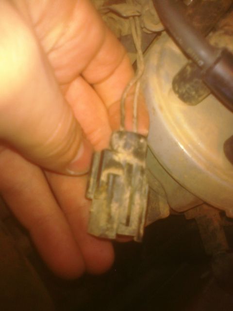 1995 Ford Bronco - A hand holds a dirty, corroded multi-pin electrical connector/plug with attached wiring, held up in front of a grimy round component (possi…